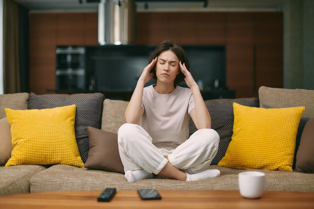 Eine Dame sitzt auf ihrem Sofa und hält sich den Kopf, weil sie Kopfschmerzen hat